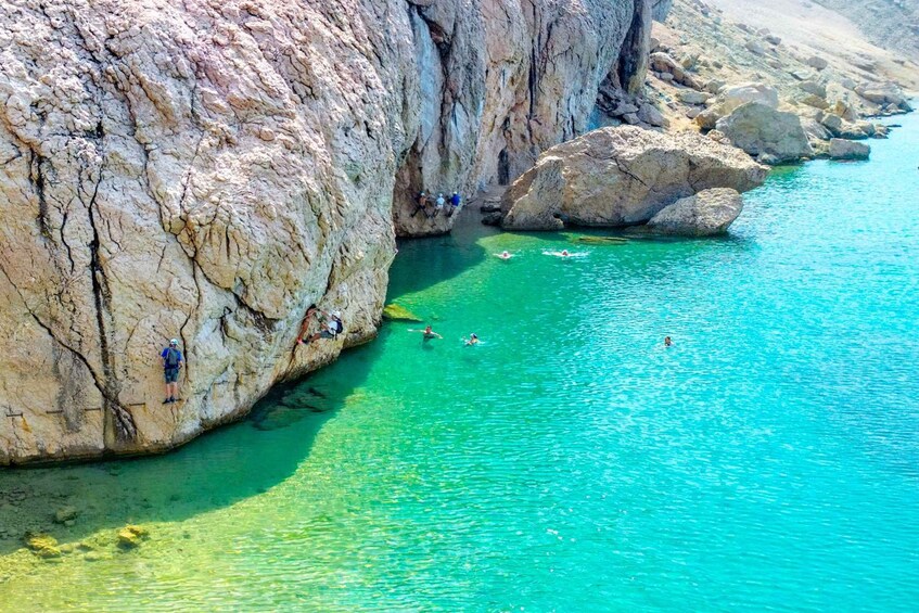 A rocky cliff with clear turquoise water, people swimming and climbing. 3