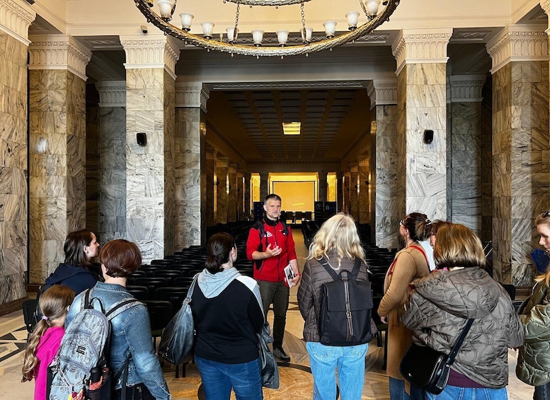 A group of people standing in a marble-floored corridor with columns, a chandelier, and a speaker addressing the crowd. 1