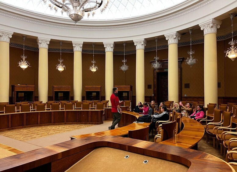 A formal meeting room with a speaker addressing an audience seated around a U-shaped table. 3