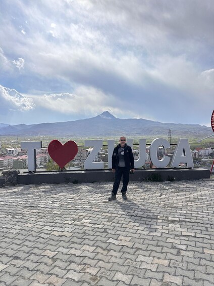 A person standing in front of a sign with the text 'I ♥ ZUCA' and a mountain in the background. 2
