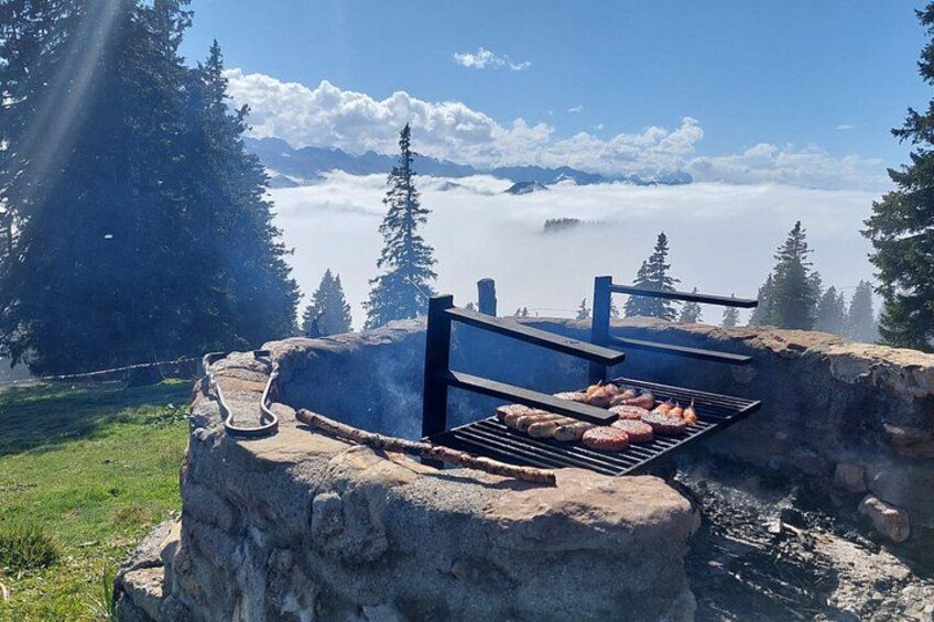 A stone structure with a grill and a hanging rack, surrounded by a mountainous landscape with trees and a clear sky. 1