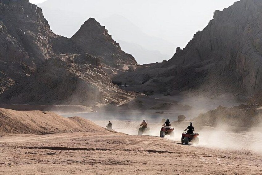 A group of people riding off-road vehicles in a desert landscape with rocky terrain and a body of water. 3