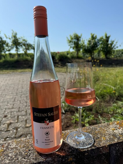 A bottle of Stefan Sauer Franken rosé wine and a glass of rosé wine on a stone ledge with greenery in the background. 1