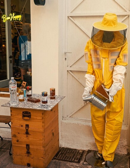 A person in a yellow bee suit and hat standing next to a wooden box with jars of honey and a water bottle. 3