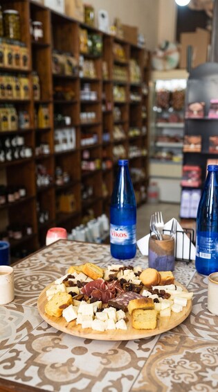 A cheese platter on a patterned tablecloth with various cheeses, crackers, and a bottle of Vikos water. 4