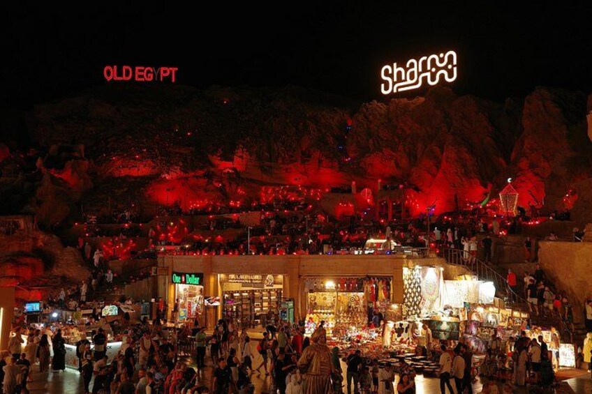 Un mercado al aire libre concurrido de noche, con letreros iluminados y un telón de fondo rocoso. 4