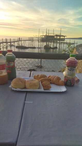 A picnic table with croissants, a bottle of juice, and a bottle of yogurt by a waterfront with a pier in the background. 2