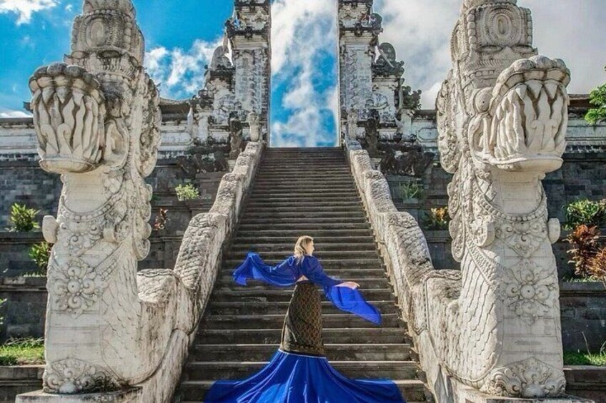 Une personne vêtue d’une robe bleue se tient au sommet d’un escalier en pierre orné de sculptures complexes, qui mène à une structure ressemblant à un temple. 3