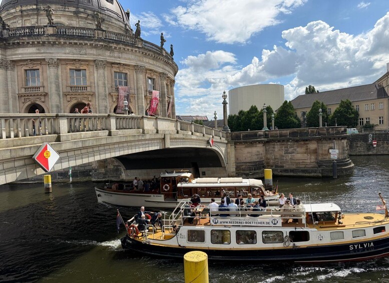 A boat named SYLVIA on a river, a bridge with people, and a historic building with flags. 1