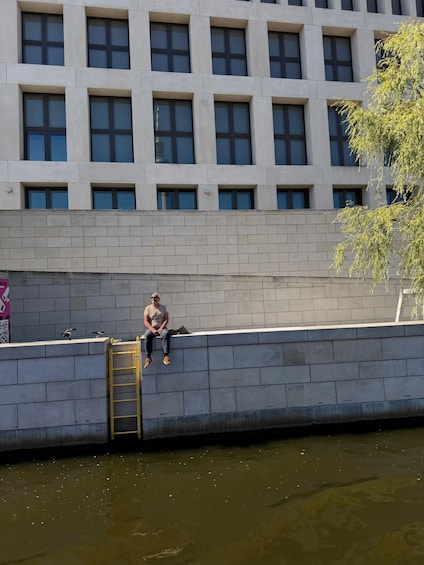 A man sitting on a ledge by a river, with a building in the background. 2