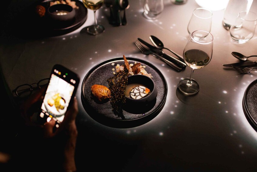 A person taking a photo of a dish with a bowl, a glass of wine, and cutlery on a table. 3