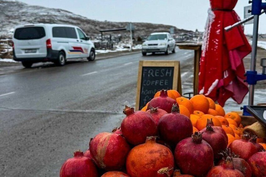 Un puesto de frutas al borde de la carretera con granadas y naranjas, un letrero que anuncia café fresco, y una camioneta estacionada en la vía. 5
