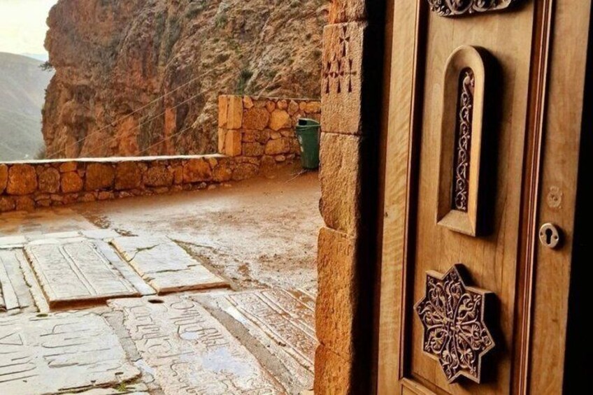 Un muro de piedra, una puerta de madera con tallas intrincadas y un paisaje rocoso. 2
