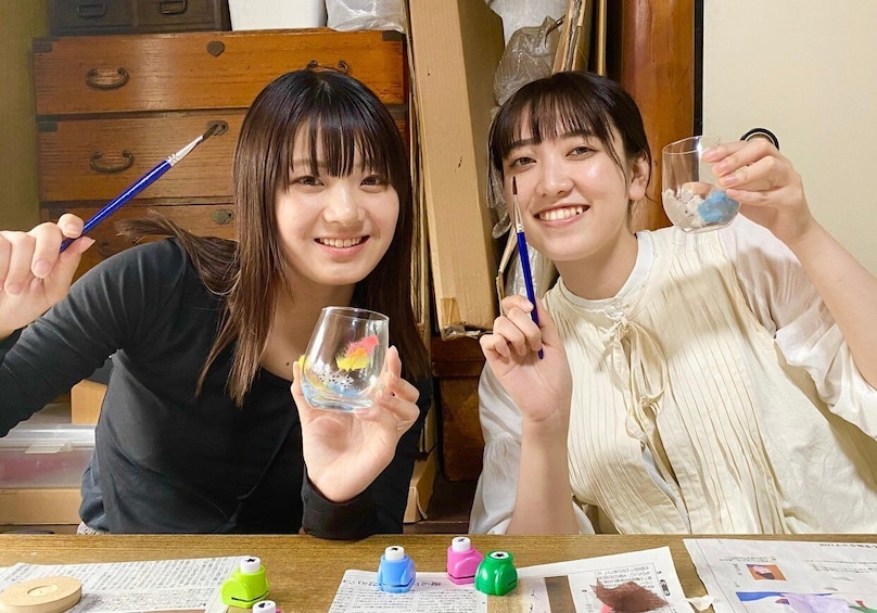 Two people holding glasses with paintbrushes, a wooden dresser, and papers on a table. 3