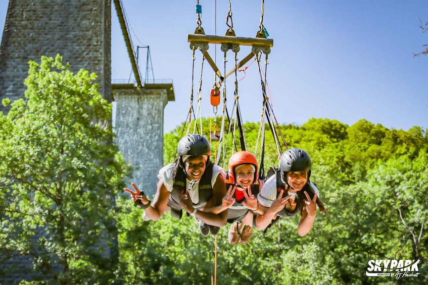 Three people on a zip line with a stone building and trees in the background. 1