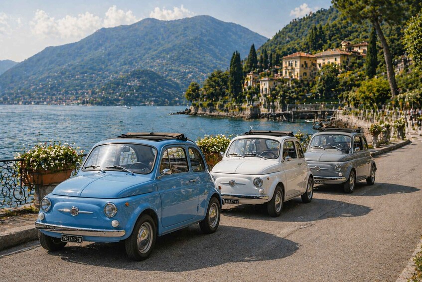 Three vintage cars parked by a lakeside road with mountains in the background. 2