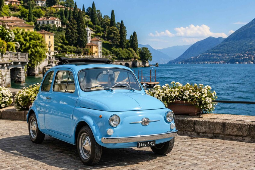 A blue vintage car parked by a lakeside with buildings and mountains in the background. 3