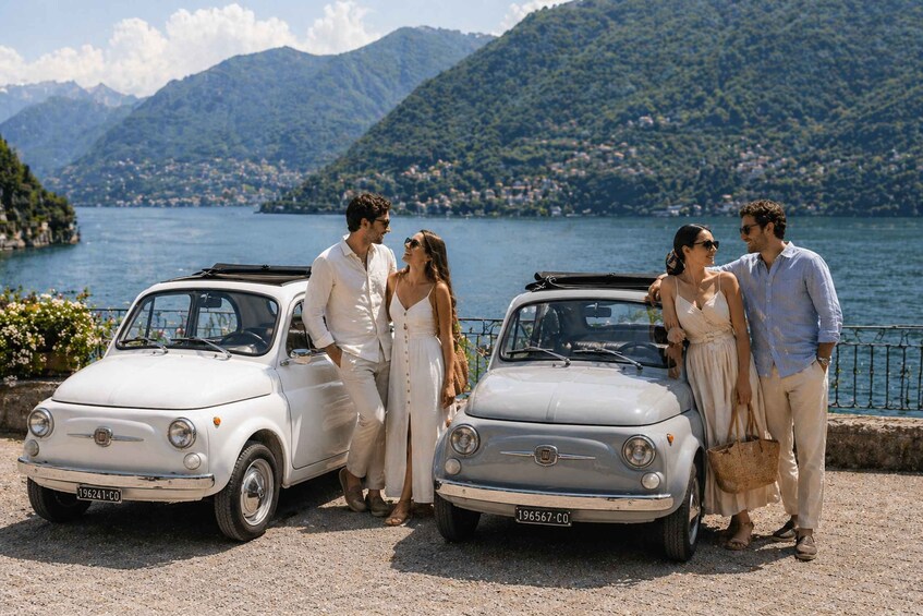 Two vintage cars parked by a lakeside with mountains in the background. 1
