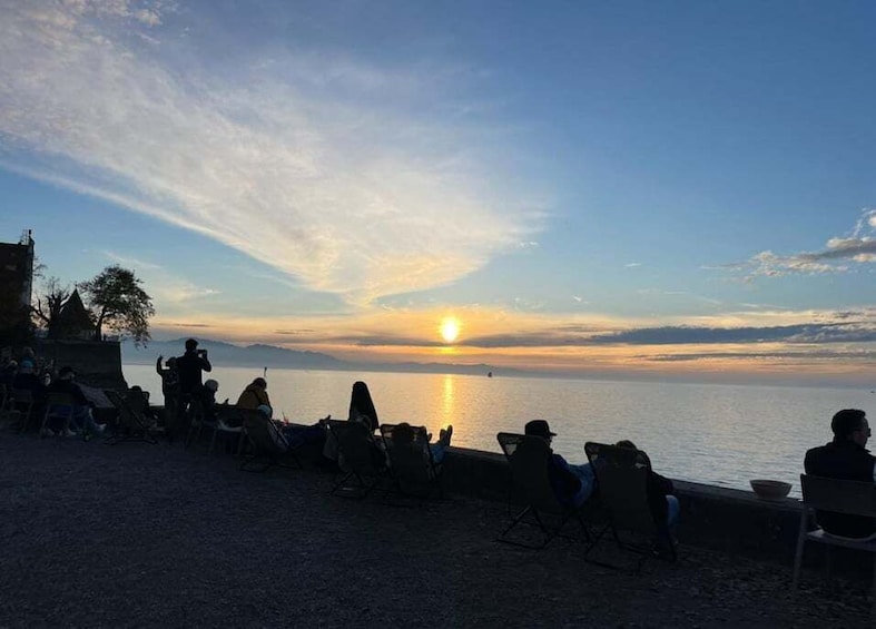 A sunset over a calm sea with people sitting on chairs along the shore. 1