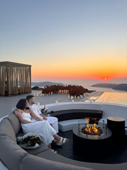 A couple sitting on a sofa overlooking a sunset, with a decorative 'MARRY' sign and candles in the background. 3