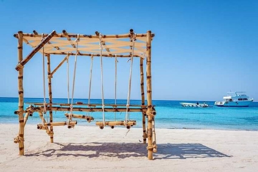 Un conjunto de columpios de bambú sobre una playa arenosa, con agua azul cristalina y barcos al fondo. 1