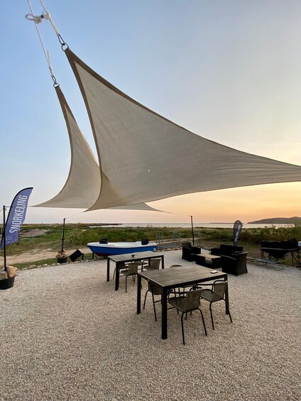 A beachside dining area with tables and chairs, a blue boat, and a large sail shade. 5