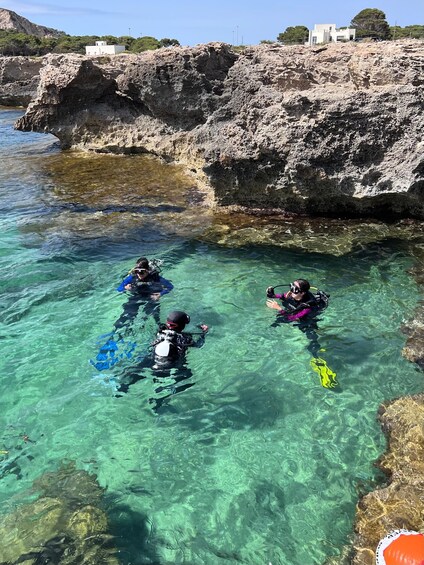 Three divers in clear water near rocky cliffs with buildings in the background. 1