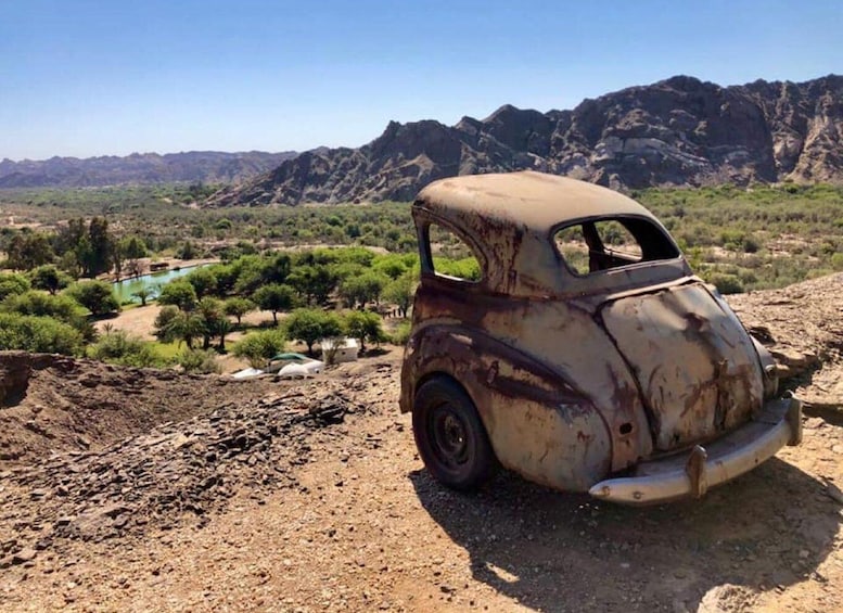 An old, rusted car parked on a rocky terrain with a mountainous backdrop and a green landscape with trees and a pond. 1