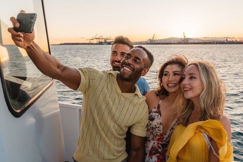 A group of people on a boat taking a selfie with a sunset and industrial structures in the background. 4