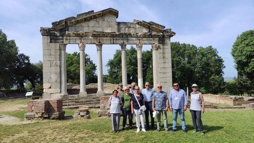 A group of people standing in front of ancient Greek ruins with columns and a partially intact temple structure. 1