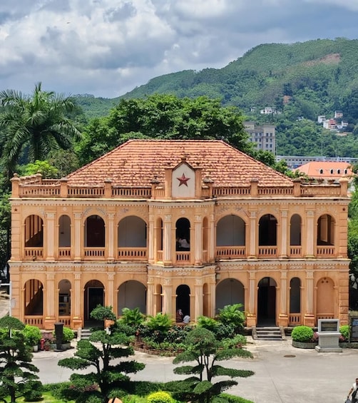 A historic building with a red star emblem, surrounded by lush greenery and a mountainous backdrop. 1