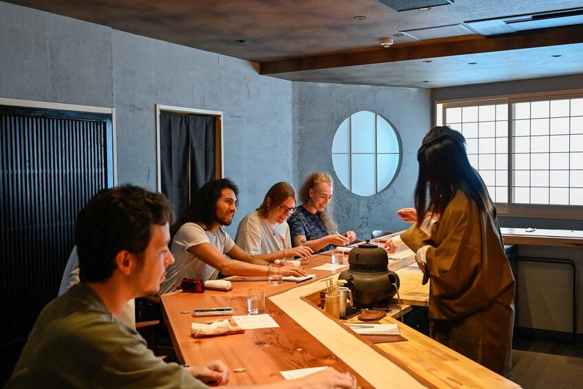 A group of people seated around a wooden table in a room with a large window and a round mirror on the wall. 1