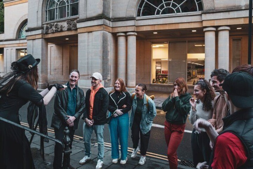 A group of people standing on a sidewalk in front of a building with large windows and columns. 3