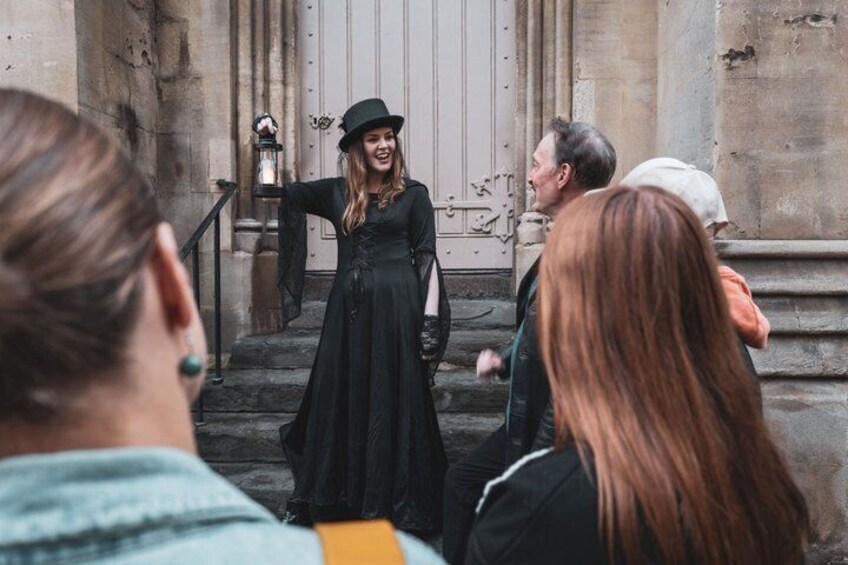 A person in a long black dress and hat standing on steps, gesturing to an audience. 5