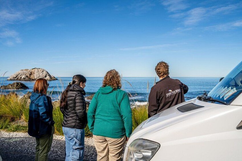 Three people standing by a white car, overlooking a coastal area with rocks and the ocean. 2