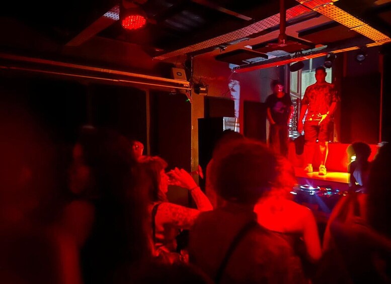 A dimly lit indoor venue with a stage, red lighting, and a crowd of people. 2
