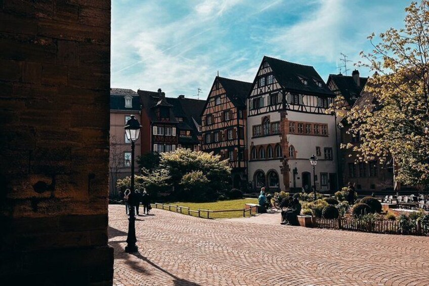 Ein Kopfsteinpflasterplatz mit traditionellen Fachwerkhäusern, einem Brunnen und einem strahlend blauen Himmel. 4
