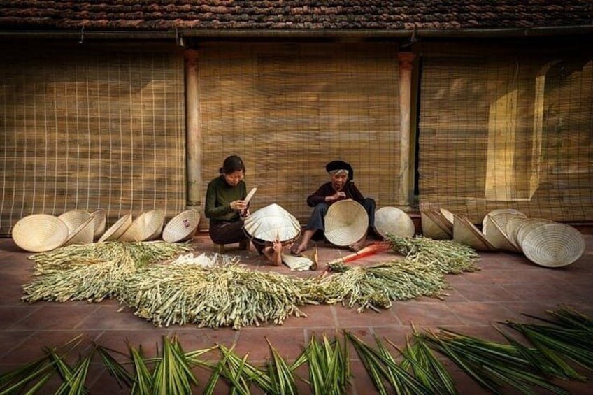 Two people sitting on the ground surrounded by large woven baskets and bundles of green plants. 3