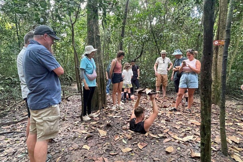 A group of people in a forested area, some standing and others sitting on the ground, engaged in an outdoor activity. 3