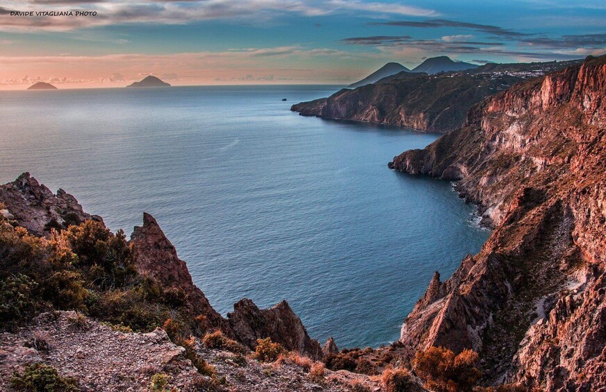 A coastal landscape with rugged cliffs, a calm sea, and distant mountains. 2