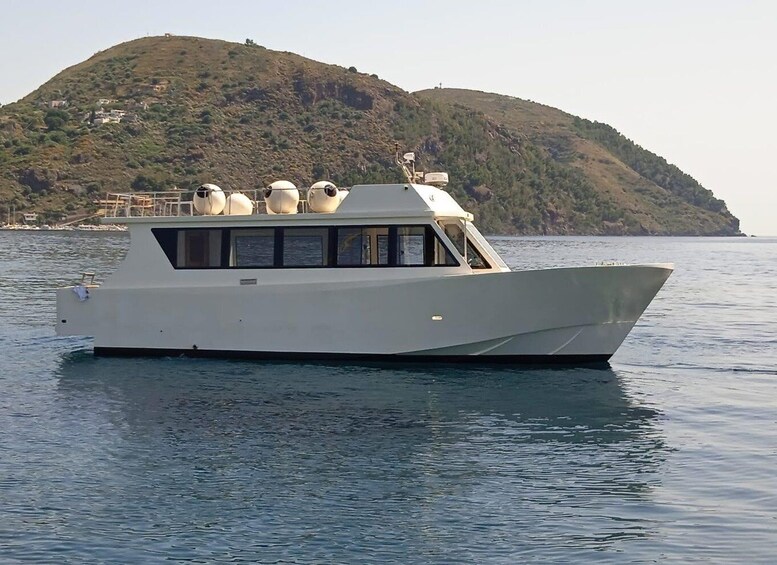 A white boat with a cabin and several spherical objects on its deck, sailing on calm waters with a hilly island in the background. 1