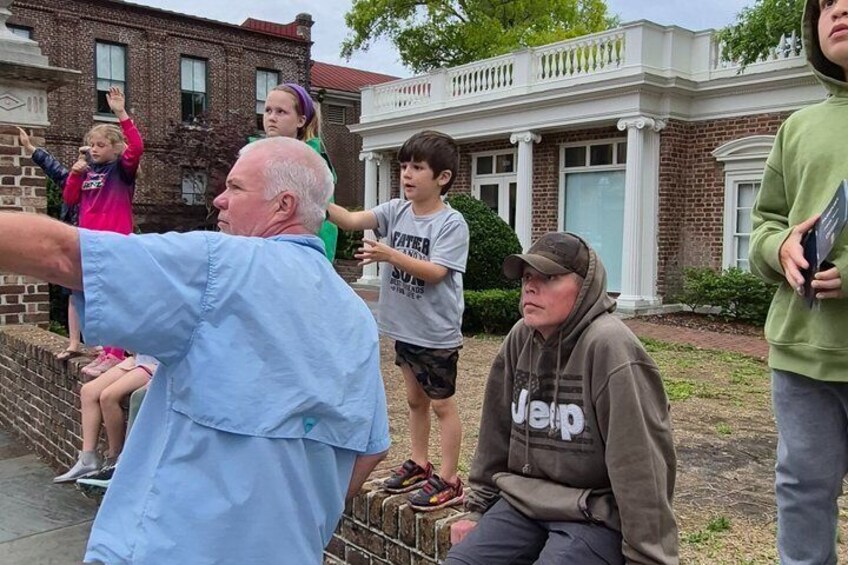 A group of people sitting and standing on a sidewalk in front of a brick house with a white porch. 2