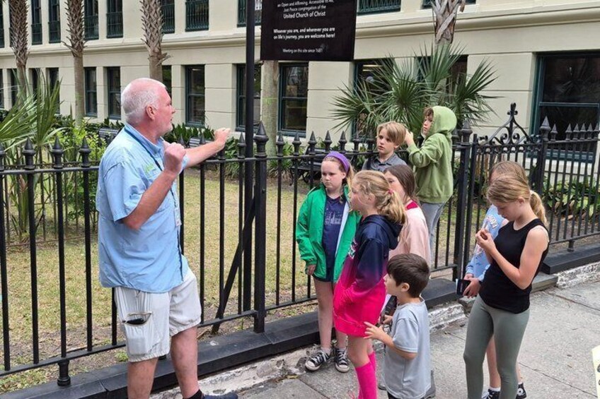 A group of people standing by a fence, a building with a sign, and a palm tree in the background. 3