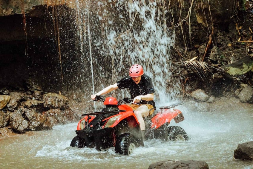 A person riding an ATV through a shallow water crossing with a waterfall in the background. 1