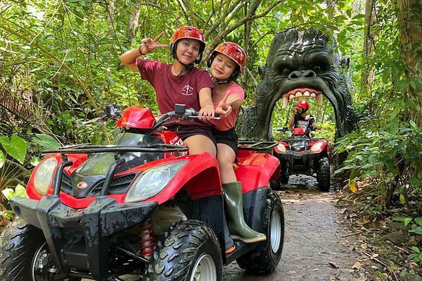 Two people on a red ATV in a forested area with a large, menacing-looking statue in the background. 2