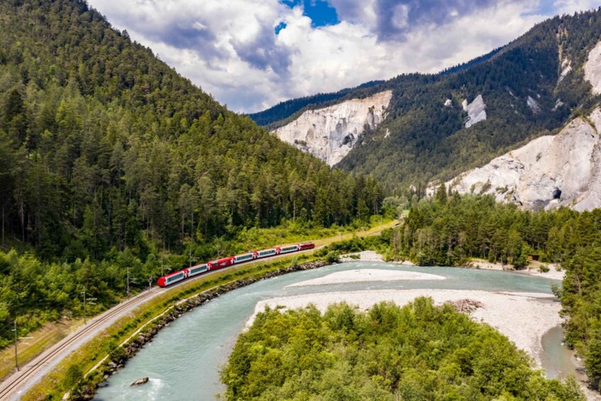 A red train traveling along a curved track beside a river, surrounded by dense forest and rocky mountains. 1