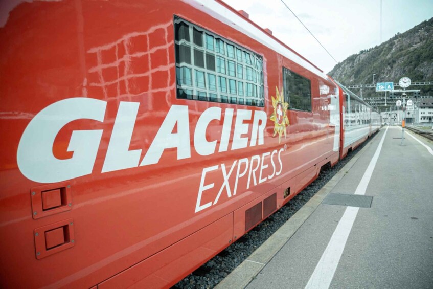 A red Glacier Express train with 'GLACIER EXPRESS' written on its side, parked at a station with a mountainous backdrop. 4