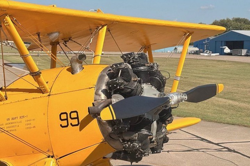 A yellow biplane with the number 99 on its side, parked on a tarmac with a grassy field and hangars in the background. 5