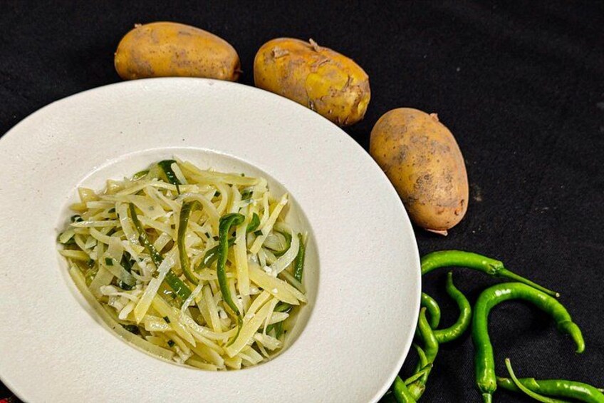 A plate of sliced vegetables with green peppers, surrounded by whole potatoes and green chili peppers on a dark surface. 3