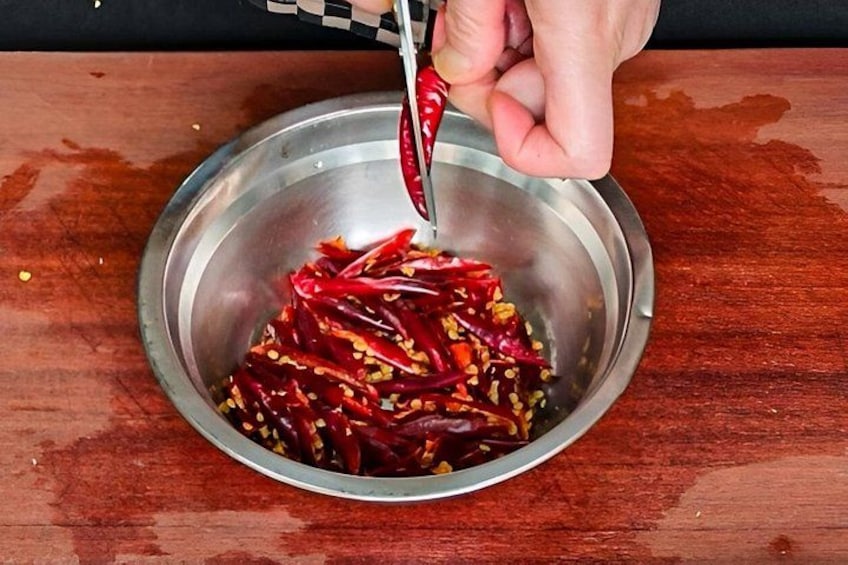 A hand holding a red chili pepper over a metal bowl containing chopped red chilies. 5
